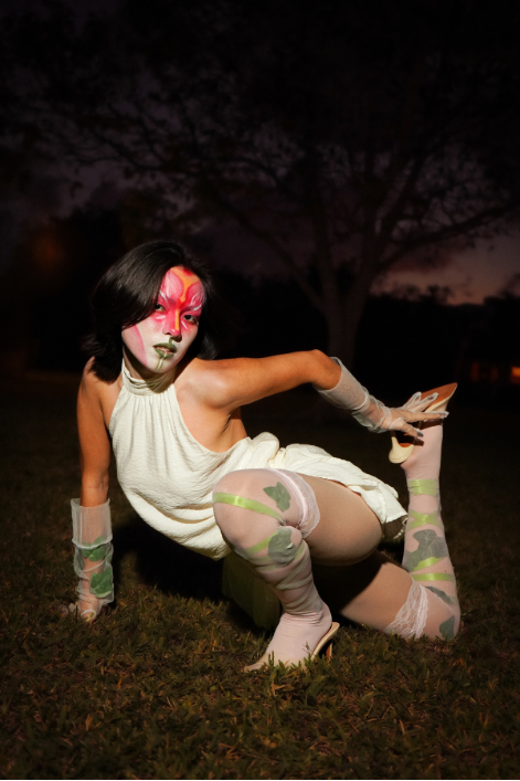 Model kneeling on grass at night in a white dress and printed tights, with pink face makeup