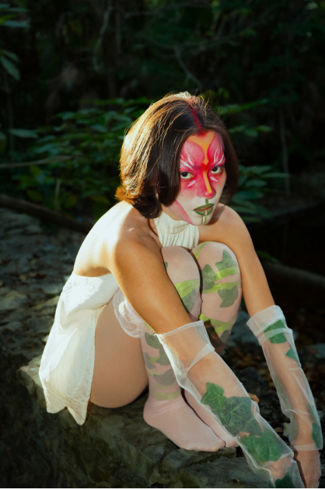Model seated on a rock in a white dress and ivy-printed gloves, with pink face makeup