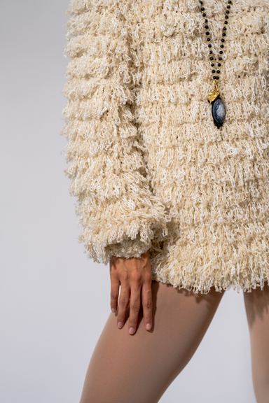 Close-up of a cream shag jacket, a pendant necklace, and a hand resting on the thigh